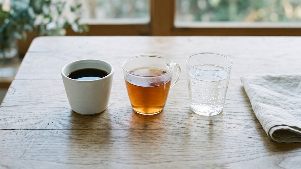 A cup of black coffee, a glass cup of tea and a glass of sparkling water in a row on a wooden table, beside a folded linen napkin.