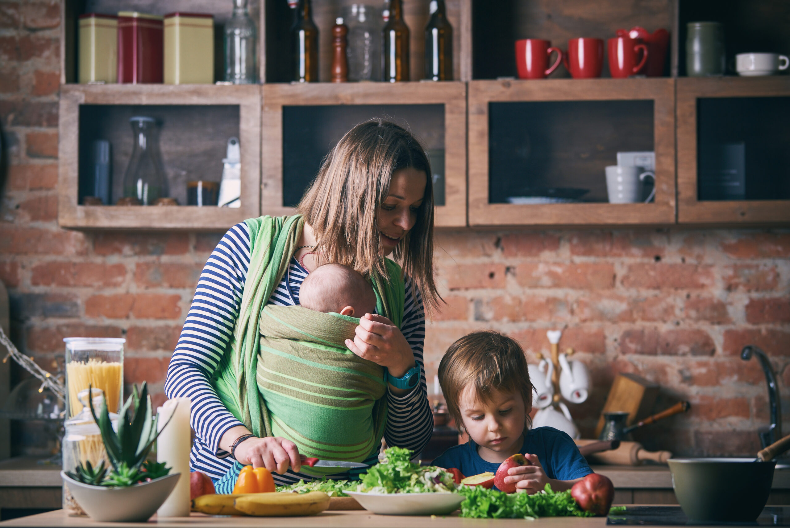 Happy young family, beautiful mother with two children, adorable preschool boy and baby in sling cooking together in a sunny kitchen.