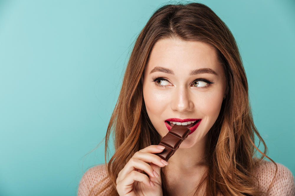 eating chocolate for brain health Portrait of a smiling brown haired woman with bright makeup eating chocolate bar isolated over blue background