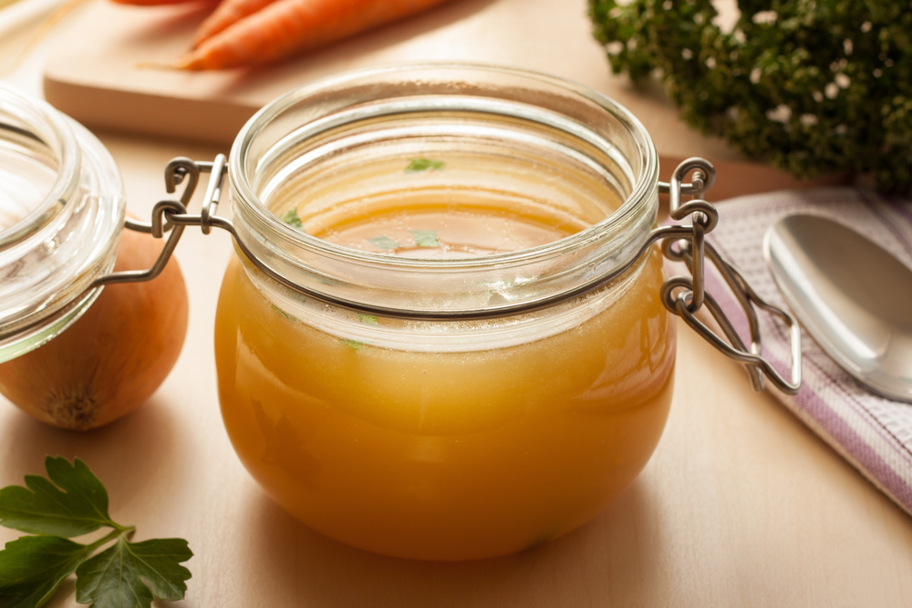 DIY Bone Broth Bone broth made from chicken in a glass jar, with carrots, onions, and parsley in the background