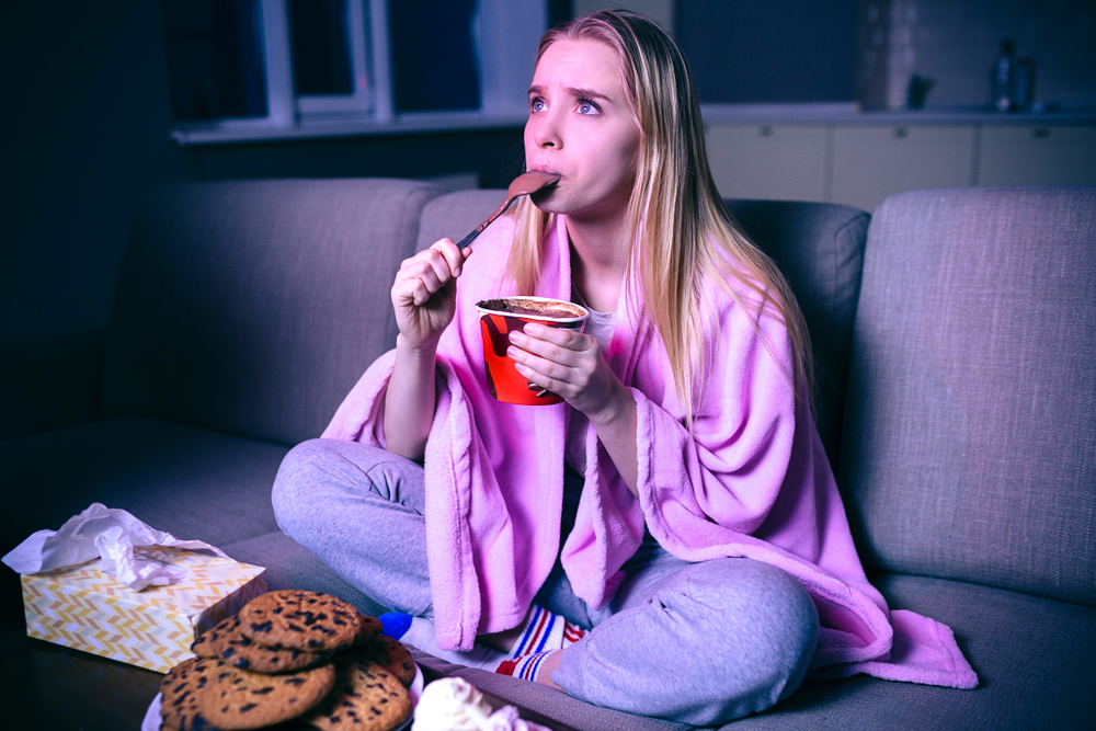 snacking late at night is bad for your health and work productivity Young woman watching movie at night. Concentrated model eating ice cream with spoon. Sitting on sofa in room alone. Streaming show or tv series.