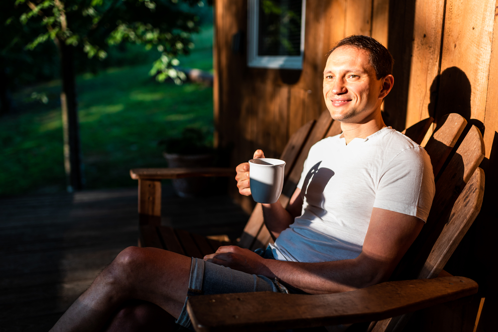 feel good while fasting Man happy sitting relaxing on rocking chair lounge on porch of house in morning wooden cabin cottage drinking coffee or tea from cup mug